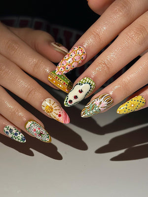 A set of long, almond-shaped fingernails, each painted with a unique design. The designs include floral patterns, suns, and geometric shapes in a variety of colors, including pink, yellow, green, and white. The nails are well-manicured and showcase i