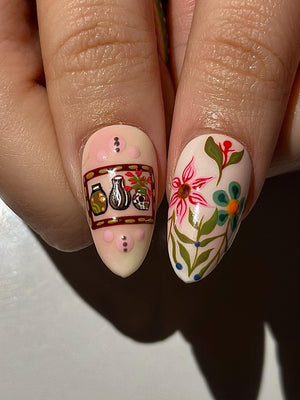 Close-up of two fingernails with detailed nail art. One nail features a design with jars, a vase, and a skull. The other nail showcases a floral design with pink and blue flowers and green leaves. The nails are painted on a light pink base.