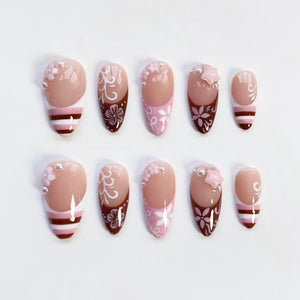 A set of ten artificial nails in various designs. The nails feature a nude base with accents of pink, white, and brown. Some nails have floral patterns, pearls, or star-shaped embellishments. Others have striped or swirl designs.