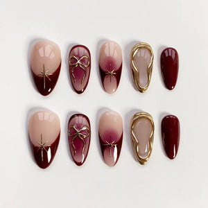 A set of ten artificial nails in various designs. Some nails are nude with a dark red tip and gold starburst, others feature a gold bow, and some have a gold frame. The nails are oval-shaped and have a glossy finish.