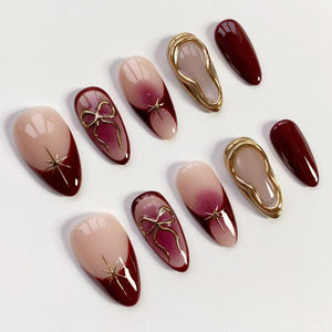 A collection of artificial nails in various designs. Some nails feature a nude base with a deep red tip and gold starburst accents. Others have a gradient effect, gold bow details, and gold-framed shapes.