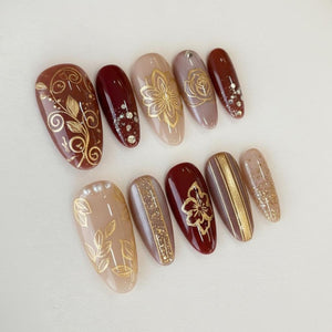 A set of ten artificial nails in various shades of brown, beige, and burgundy. Each nail features gold floral or geometric designs, including leaves, flowers, and stripes. Some nails have glitter or small gem accents.