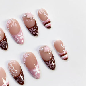A collection of oval-shaped false nails. The nails feature a variety of designs, including floral patterns, star-shaped embellishments, and striped accents. The color palette includes shades of pink, brown, and white, creating a delicate and feminine