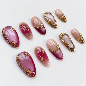 A collection of oval-shaped artificial nails in various designs. Some nails feature a gradient of pink and burgundy, adorned with gold embellishments and rhinestones. Others have a gold frame with a white interior, and some are a solid nude color.