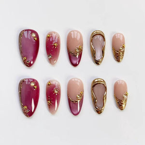 A set of ten artificial nails in various designs. Some nails are a deep burgundy with gold accents, while others are a nude pink with gold embellishments. One design features a gold frame.