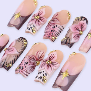 A collection of artificial nails with a floral design. The nails feature a nude base with a dark brown tip and are adorned with pink flowers, gold accents, and animal print details. Some nails have a gold star design.