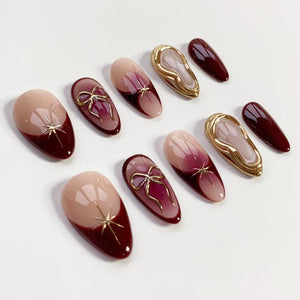 A collection of almond-shaped artificial nails in various designs. Some nails feature a nude base with a deep burgundy tip and gold starburst accents. Others have a gradient effect with gold bow or star details. Additional nails are solid burgundy or