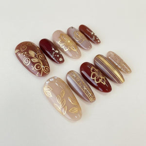 A collection of almond-shaped artificial nails in various shades of brown, burgundy, and beige. Each nail features gold foil designs, including floral patterns, leaf motifs, and glitter accents. The nails are arranged on a white surface.