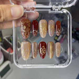 A set of ten artificial nails in a clear plastic case. The nails feature various designs, including floral patterns, glitter, and stripes, in shades of brown, gold, and beige. Some nails have rhinestones. The nails are oval-shaped.