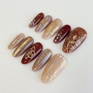 A collection of ten artificial nails in various shapes and colors. The nails feature designs including gold floral patterns, glitter, and stripes. Colors include burgundy, beige, and gold, with some nails adorned with small rhinestones.