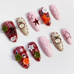 A collection of almond-shaped false nails with various designs. Some nails feature a red plaid pattern, while others are solid pink with white polka dots. Embellishments include star and apple designs, buttons, and a small orange teddy bear.