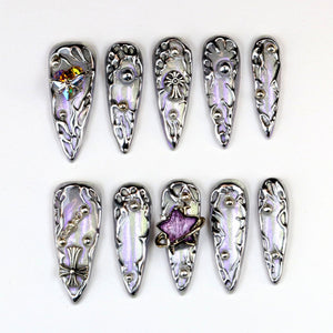 A set of ten long, silver-toned artificial nails. Each nail features a pearlescent, iridescent finish with ornate, raised silver designs and decorative embellishments, including gems and crosses. The nails are arranged on a white surface.