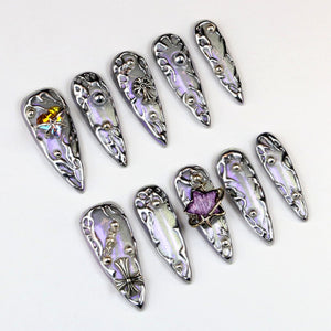 A collection of ten silver-toned, ornate nail art designs. Each nail features a pearlescent lavender base with intricate metal embellishments, including crosses, stars, and decorative studs. The nails are arranged on a white surface.