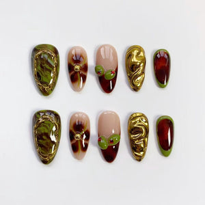 A set of ten artificial nails in various designs. The nails feature olive green, gold, and burgundy colors. Some nails have gold embellishments, while others have olive and red accents. The nails are oval-shaped.