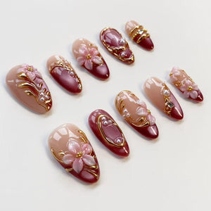 A collection of artificial nails in various designs. The nails feature a gradient of nude and burgundy colors, adorned with gold accents, pearls, and 3D floral embellishments. Some nails have a gold border, while others have a pearl accent.