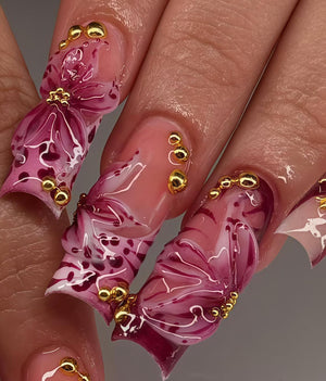 Close-up of long, artificial nails with a floral design. The nails are painted with a gradient of pink and white, adorned with gold beads. The floral design is a deep burgundy color.