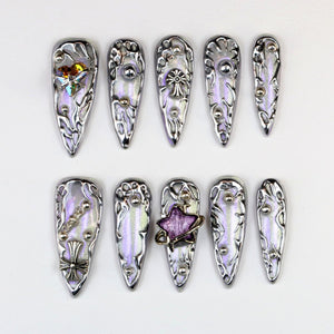 Set of ten silver and iridescent false nails. Each nail features a unique design with decorative elements like studs, crosses, and a star. The nails have a pointed shape and are arranged on a white surface.