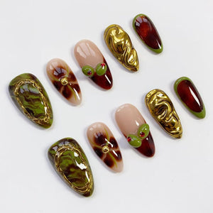 A collection of artificial nails in various designs. Some nails feature a gold swirl design, others have a marble green and gold pattern, and some have a nude base with a dark red tip and olive accents. The nails are oval-shaped.
