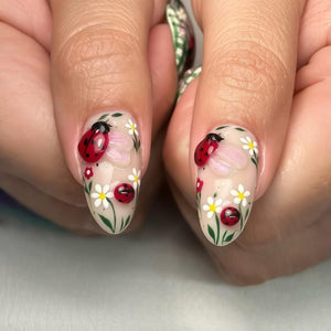 Close-up of two fingernails with detailed nail art. Each nail features a red ladybug with black spots, white daisies, pink flowers, and green leaves on a clear base. The design is intricate and nature-themed.