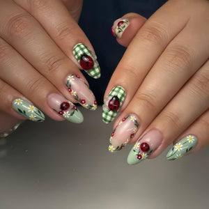 A set of long, oval-shaped nails with detailed nail art. Designs include ladybugs, daisies, and green gingham patterns. The nails are painted in shades of green, pink, and white, creating a spring-like aesthetic.