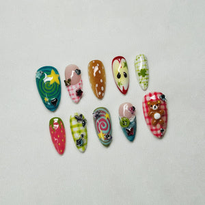 A set of ten artificial nails with various designs. The nails feature a range of colors, including teal, pink, brown, red, and green. Designs include a star, polka dots, plaid, an apple, a teddy bear, and swirls, with added embellishments.