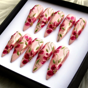Set of ten long, almond-shaped artificial nails. Each nail is painted with a light pink base, adorned with red floral designs, green leaves, and gold accents. The nails are arranged in a black box.