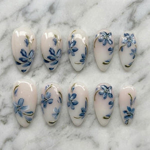 A set of ten oval-shaped artificial nails. Each nail is painted with a light pink base and features a blue floral design with gold accents. The nails are arranged on a white marble surface.