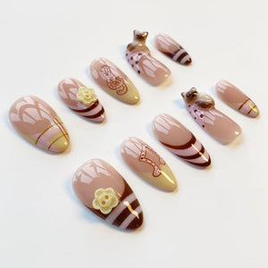 A set of ten artificial nails in various designs. The nails feature a light pink base with brown and beige accents. Some nails have floral or musical note designs, while others have cat-shaped embellishments.