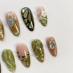 A collection of artificial nails with various designs. Some nails feature gold embellishments, iridescent stones, and floral accents. Others showcase a butterfly wing pattern or a swirl design in shades of green and pink.