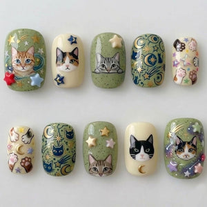 A set of ten artificial nails with cat-themed designs. The nails feature various cat faces, stars, moons, and paw prints in shades of green, cream, and gold. Some nails have 3D star accents.