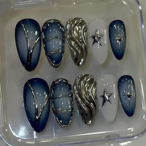 A set of ten artificial nails in a clear plastic container. The nails feature a variety of designs, including a gradient blue and silver, silver wave, white with a silver star, and a blue gradient with a silver starburst.