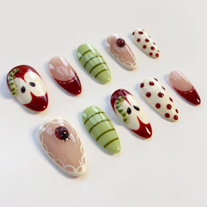 A collection of ten artificial nails with various designs. The nails feature a variety of colors, including red, green, pink, and white. Some nails have apple, ladybug, and striped patterns. Others have a solid color or a French tip design.