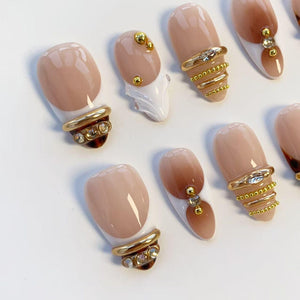 A set of artificial nails in various designs. Each nail features a nude base with a French tip or gradient effect. Some nails have gold rings with rhinestones or small gold beads for added embellishment. The nails are arranged on a white surface.