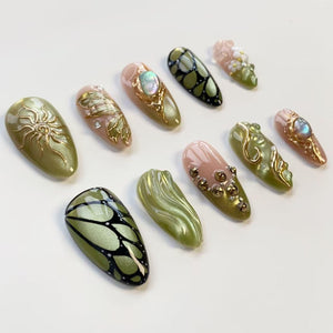 A collection of ten almond-shaped, decorated artificial nails. Designs include gold sunbursts, butterfly patterns, and floral accents. Colors range from olive green and pink to gold and black, with iridescent and metallic finishes.