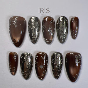 A set of ten artificial nails in various shapes, featuring a metallic silver and brown color scheme. Each nail is adorned with intricate, embossed floral designs. Some nails have a central jewel accent. The brand name "IRIS NAILAVY" is visible.