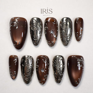 A set of ten artificial nails in a variety of shapes. The nails feature a metallic silver and brown color scheme with ornate, swirling designs. Some nails have a central jewel. The brand name "IRIS NAILXY" is at the top.