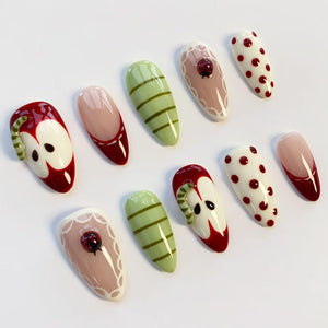 A collection of ten artificial nails with various designs. The nails feature designs such as red apples, green stripes, red polka dots, and a ladybug. Some nails have a nude base with a red tip or white lace.