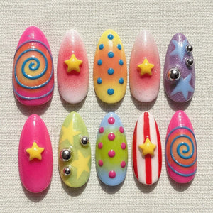 A set of ten artificial nails in various designs. The nails feature bright colors like pink, yellow, and purple, with embellishments such as stars, swirls, dots, and metallic accents. The nails are oval-shaped.