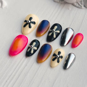 A collection of artificial nails in various designs. Some nails are cream-colored with black floral designs, others are chrome, and some feature gradient colors of pink and orange or blue and brown. A small silver gem is at the center of some of the