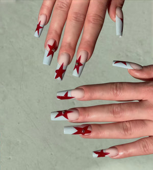 Long, square-shaped artificial nails with a light blue base and red star designs. Some nails feature a small, silver gem at the star's center. The nails have a French tip design with a clear base.