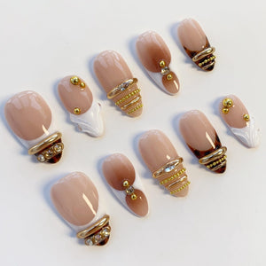 A collection of ten artificial nails with a neutral color palette. The nails feature a gradient design, gold accents, and decorative embellishments. Some nails have a white accent at the tip, while others have gold rings.