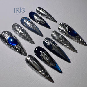 An assortment of long, pointed, silver-colored nail art designs. Some nails feature a dark blue gradient, while others have a blue gem. The Iris Nail Art logo is in the upper left corner.