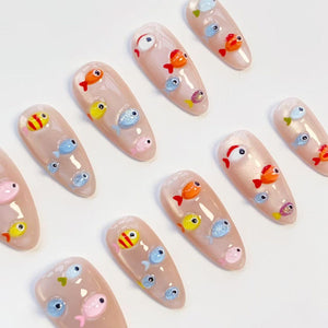 A collection of almond-shaped artificial nails with a nude base, adorned with colorful, cartoonish fish designs. The fish are in various colors, including orange, yellow, blue, and pink, with glitter accents. The nails are arranged on a white surface