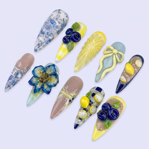 A collection of ten long, almond-shaped artificial nails. Designs include floral patterns, blueberries, lemon slices, and a bow. Colors include blue, yellow, brown, and beige. Some nails have a glossy finish, while others have a matte appearance.