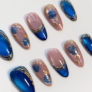 A collection of artificial nails with a variety of designs. Some nails are a solid, glossy blue, while others feature a pink base with gold accents and blue rose designs. The nails are oval-shaped.
