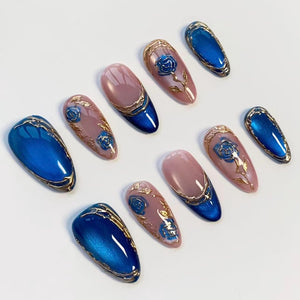 A set of ten artificial nails in various designs. Some nails are a glossy blue with gold trim, while others are a nude pink with blue and gold floral accents. The nails are oval-shaped and arranged on a white surface.