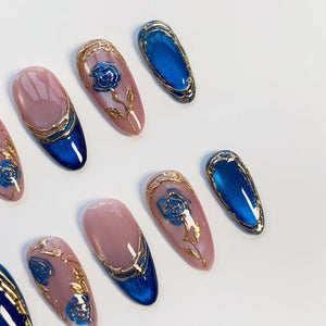 An assortment of artificial nails with a glossy finish. The nails feature a combination of pink, blue, and gold accents. Some nails have a blue rose design, while others have a solid blue color with gold trim. The nails are oval-shaped.