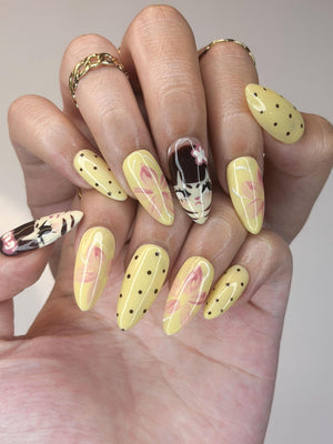 A hand displaying long, almond-shaped artificial nails. The nails are painted in a variety of designs, including yellow with black polka dots, pink floral patterns, and a detailed anime-style design. Gold rings adorn the fingers.