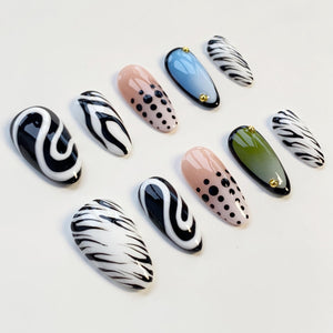 A collection of press-on nails in various designs. Some nails feature black and white swirls, zebra stripes, or black polka dots on a nude base. Others are solid colors, including blue and olive green, with gold accents.