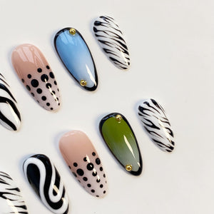 A collection of oval-shaped false nails in various designs. Some nails feature a nude base with black polka dots, while others have a blue or green gradient. Several nails display a black and white zebra stripe pattern. Each nail is adorned with a sm